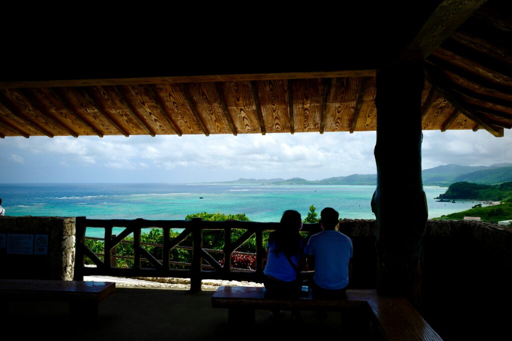 A quiet beach with crystal-clear turquoise water, traditional Japanese wooden architecture nestled among palm trees.